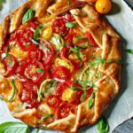 Heirloom tomato galette on a rustic wooden board, golden flaky crust folded around colorful overlapping heirloom tomato slices garnished with fresh basil and flaky sea salt