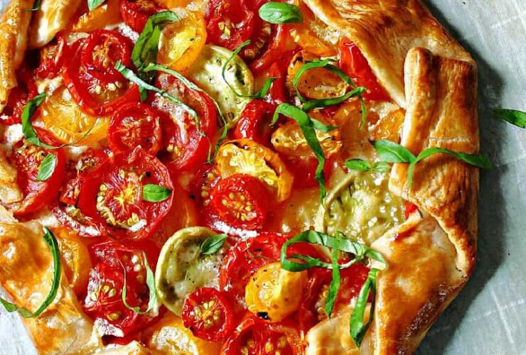 Heirloom tomato galette on a rustic wooden board, golden flaky crust folded around colorful overlapping heirloom tomato slices garnished with fresh basil and flaky sea salt