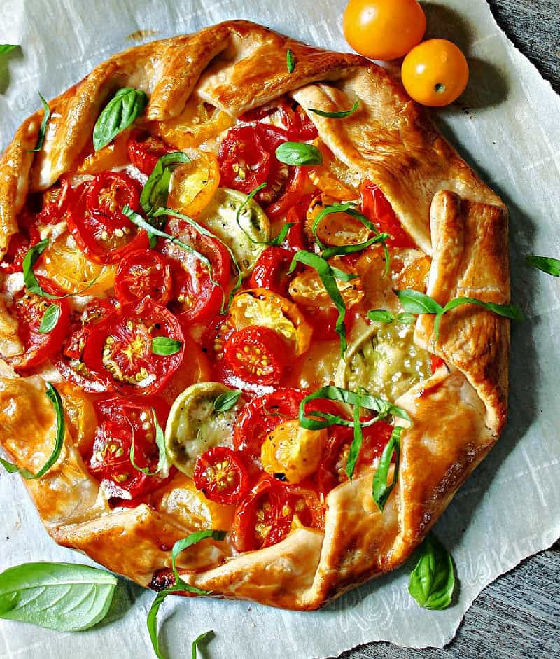 Heirloom tomato galette on a rustic wooden board, golden flaky crust folded around colorful overlapping heirloom tomato slices garnished with fresh basil and flaky sea salt