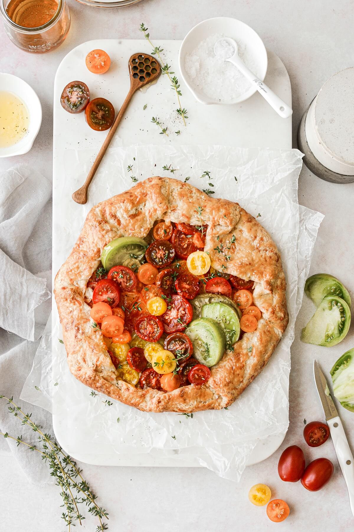 Overhead flat lay of heirloom tomato galette ingredients including sliced multicolored heirloom tomatoes, bowl of ricotta, fresh thyme sprigs, rolled pastry dough on parchment, and small bowl of olive oil on a marble surface