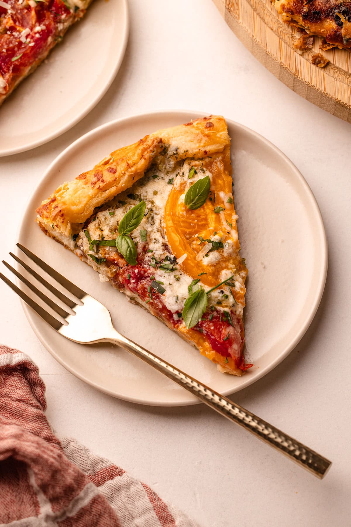 Close-up of a slice of heirloom tomato galette on a white ceramic plate showing layers of golden flaky crust, creamy ricotta, and jewel-toned roasted heirloom tomatoes topped with torn fresh basil leaves and flaky sea salt