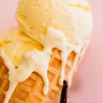 Homemade vanilla ice cream scooped into a white ceramic bowl with a golden waffle cone on the side, fresh vanilla bean pods in the background, warm natural light