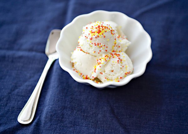 Step-by-step ingredients for homemade vanilla ice cream laid out on a white marble counter — cream, egg yolks in a glass bowl, vanilla bean, sugar, and whole milk in a small pitcher
