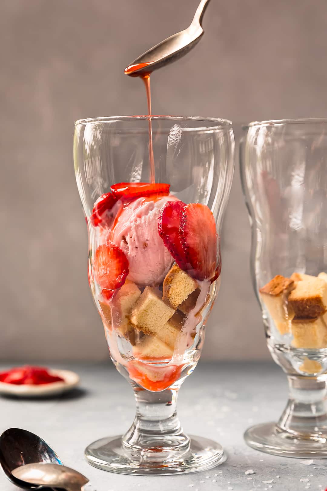 Close-up of two scoops of homemade vanilla ice cream in a vintage glass sundae dish, flecked with real vanilla bean seeds, a drizzle of honey and fresh strawberries on the side