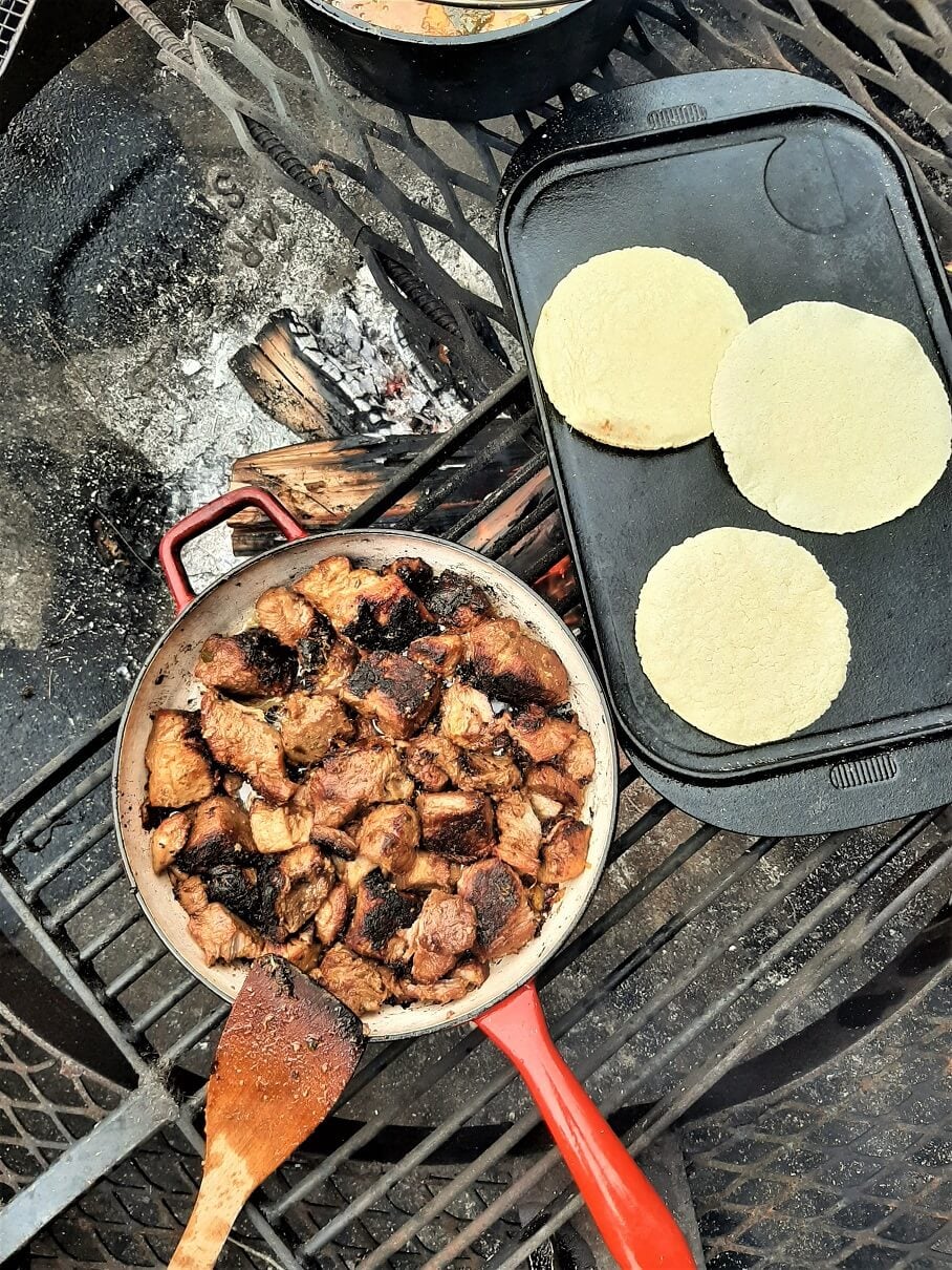 Close-up of crispy carnitas in a cast iron skillet over campfire coals, with charred edges and a drizzle of braising juices, warm corn tortillas stacked beside the pan