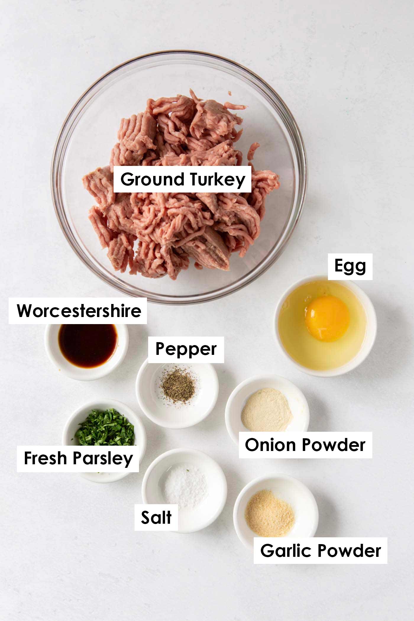 Overhead flat-lay of ground turkey, Worcestershire sauce, garlic, egg, diced onion, parsley, and spices arranged on a white marble surface ready for mixing