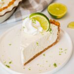 Classic Key Lime Pie with golden graham cracker crust, creamy pale yellow filling, and swirled whipped cream topping on a white plate with fresh lime slices