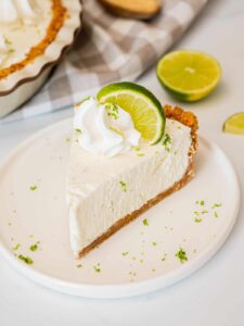 Classic Key Lime Pie with golden graham cracker crust, creamy pale yellow filling, and swirled whipped cream topping on a white plate with fresh lime slices