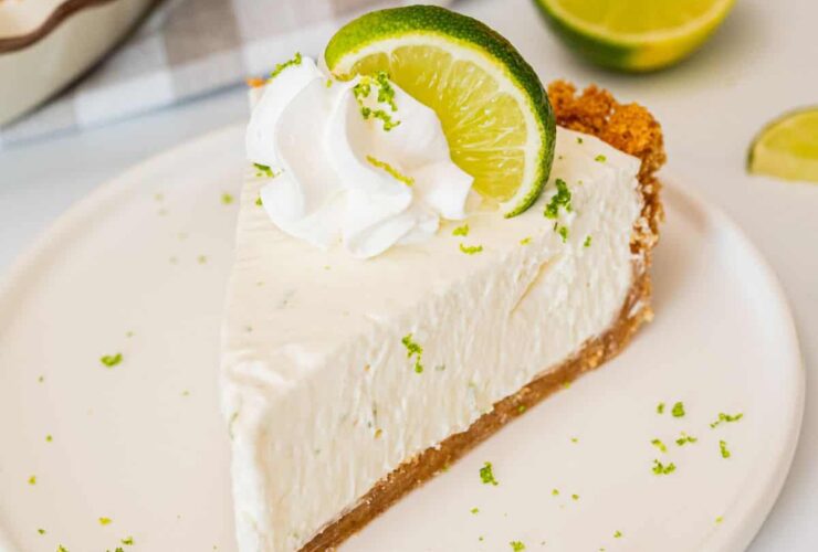Classic Key Lime Pie with golden graham cracker crust, creamy pale yellow filling, and swirled whipped cream topping on a white plate with fresh lime slices