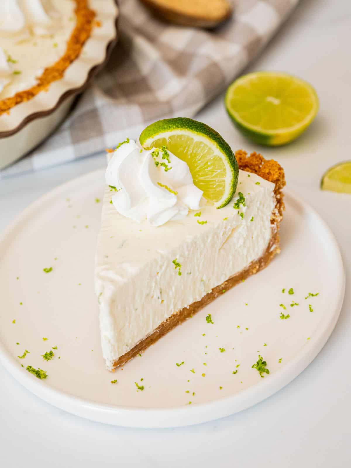 Classic Key Lime Pie with golden graham cracker crust, creamy pale yellow filling, and swirled whipped cream topping on a white plate with fresh lime slices