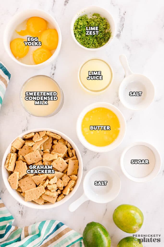 Key Lime Pie ingredients flatlay — Key limes, sweetened condensed milk cans, graham crackers, egg yolks, butter, and lime zest on a marble surface