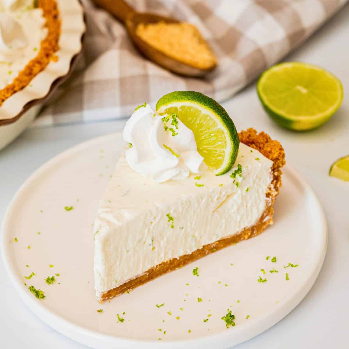 Close-up slice of Key Lime Pie on a white ceramic plate showing silky pale yellow filling, golden graham cracker crust layers, and a dollop of whipped cream with lime zest curl