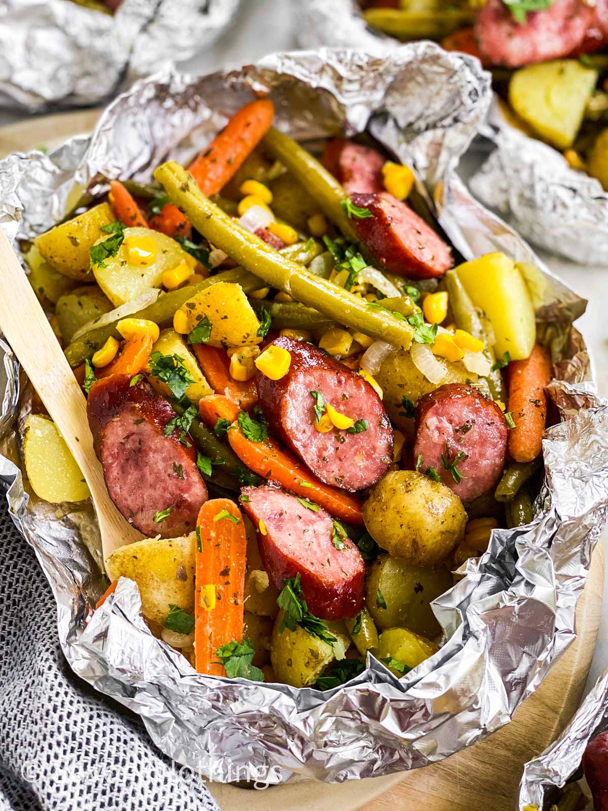 Close-up of opened kielbasa and potato foil packet showing caramelized sausage slices and golden potatoes with a glossy mustard butter sauce, served directly from the foil on a rustic wooden table