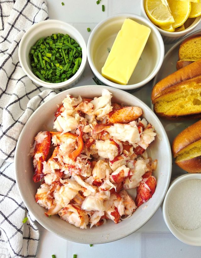 Fresh cooked lobster being shelled and chopped on a cutting board with split-top buns, butter, mayonnaise, lemon, and fresh chives arranged nearby