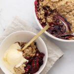 Golden mixed berry crisp with oat crumble topping in a white baking dish, surrounded by fresh strawberries, blueberries, and raspberries on a rustic wooden table