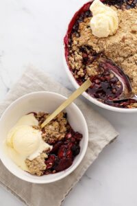 Golden mixed berry crisp with oat crumble topping in a white baking dish, surrounded by fresh strawberries, blueberries, and raspberries on a rustic wooden table