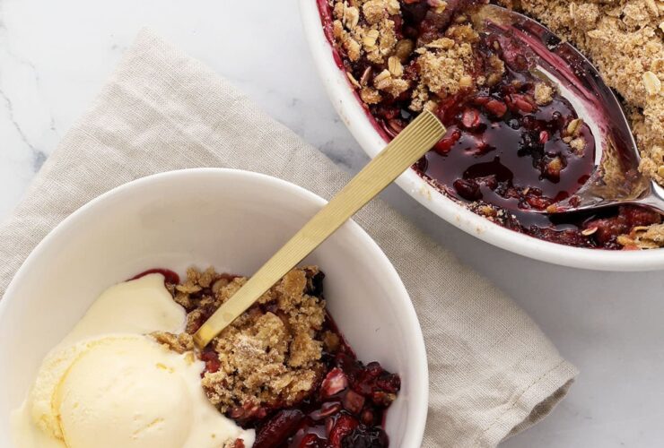 Golden mixed berry crisp with oat crumble topping in a white baking dish, surrounded by fresh strawberries, blueberries, and raspberries on a rustic wooden table