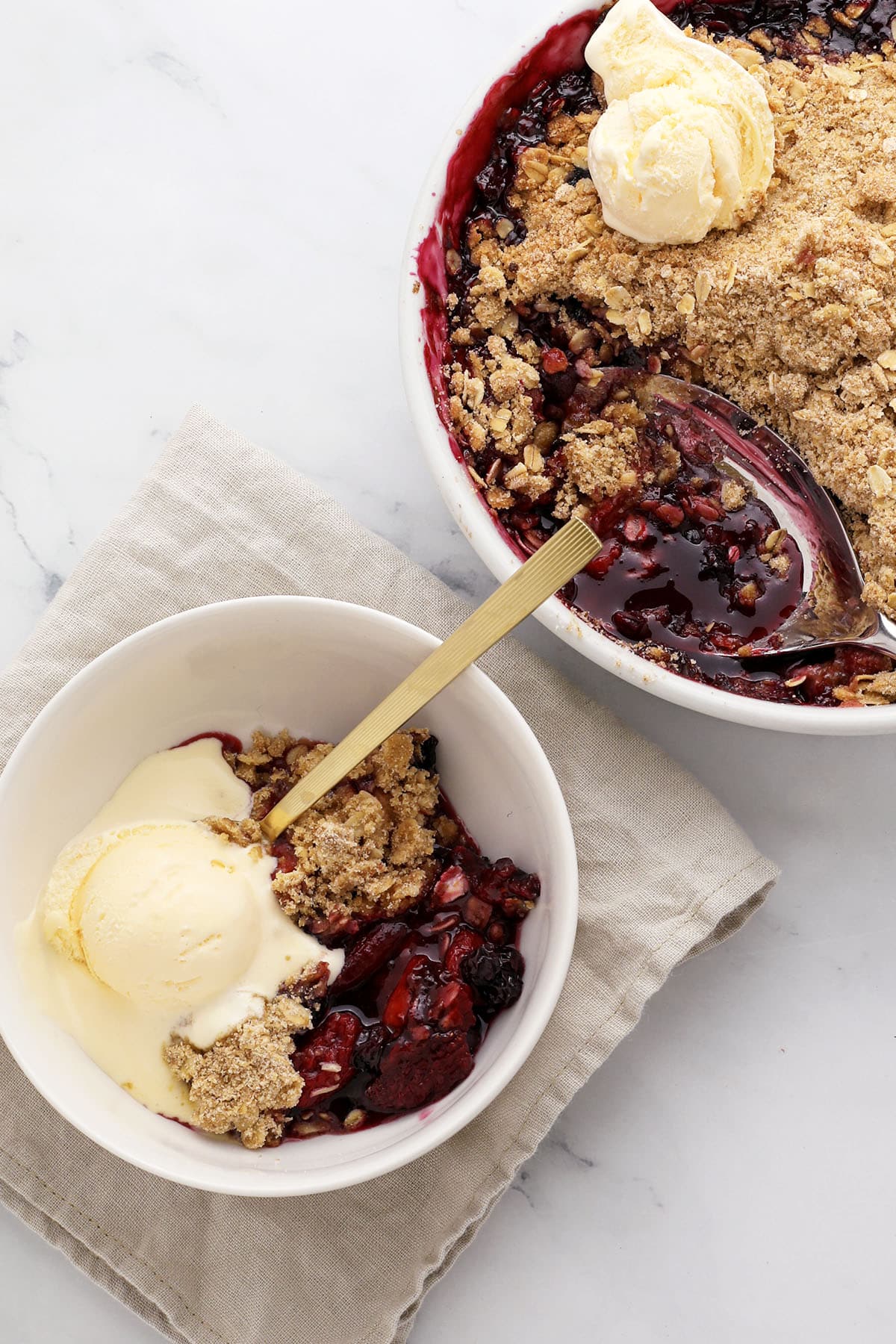 Golden mixed berry crisp with oat crumble topping in a white baking dish, surrounded by fresh strawberries, blueberries, and raspberries on a rustic wooden table