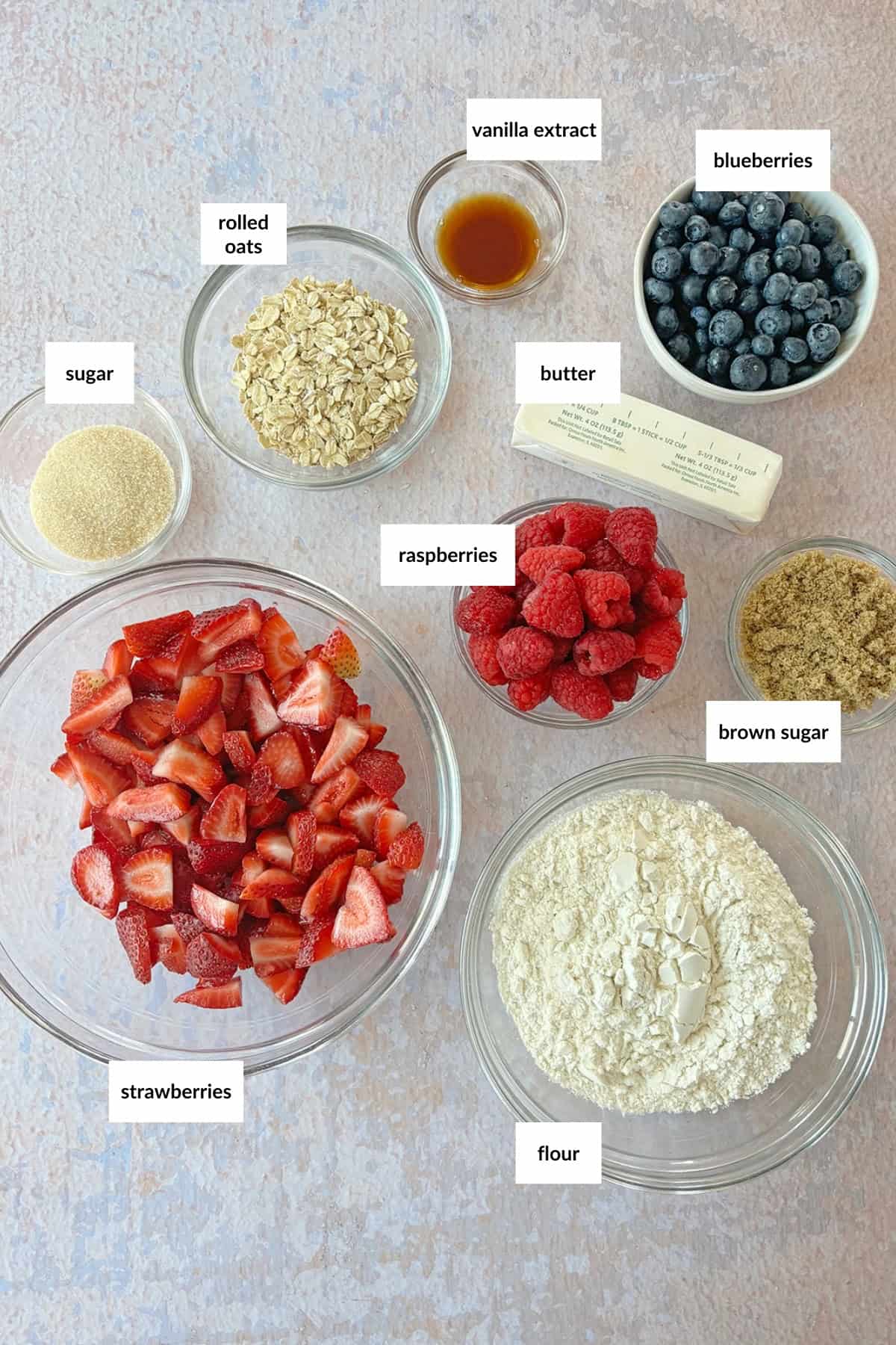 Overhead flat-lay of mixed berry crisp ingredients — rolled oats, fresh strawberries, blueberries, raspberries, maple syrup, butter, cinnamon, and brown sugar arranged on a marble surface