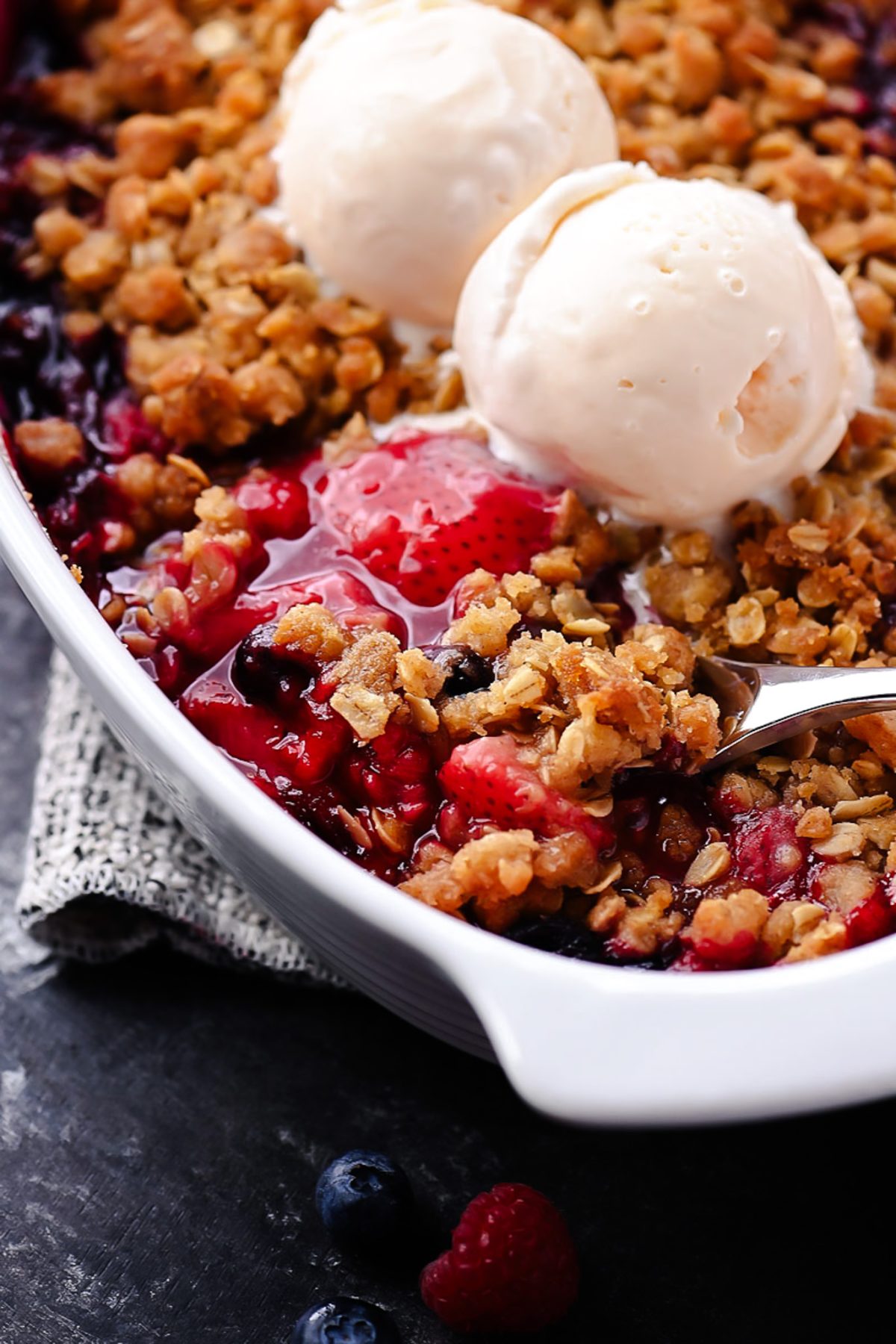 Close-up of a single serving of mixed berry crisp in a white bowl topped with a scoop of vanilla ice cream, showing the jammy berry filling and golden oat crumble topping with steam rising