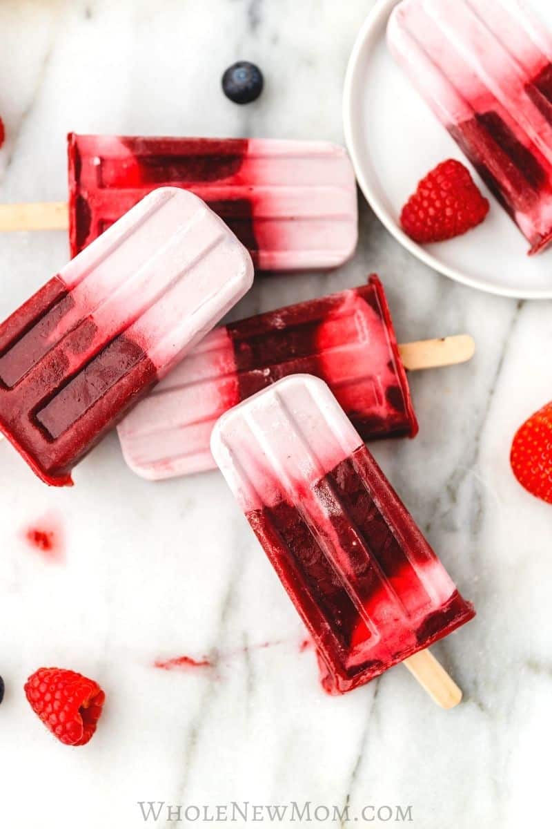 Overhead flat lay of mixed berry popsicle ingredients — fresh strawberries, blueberries, raspberries, coconut milk, honey, lemon, and vanilla on a light wooden surface