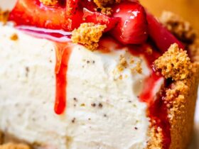 No bake cheesecake on a white cake stand with fresh strawberry topping and golden graham cracker crust, sliced to show creamy filling