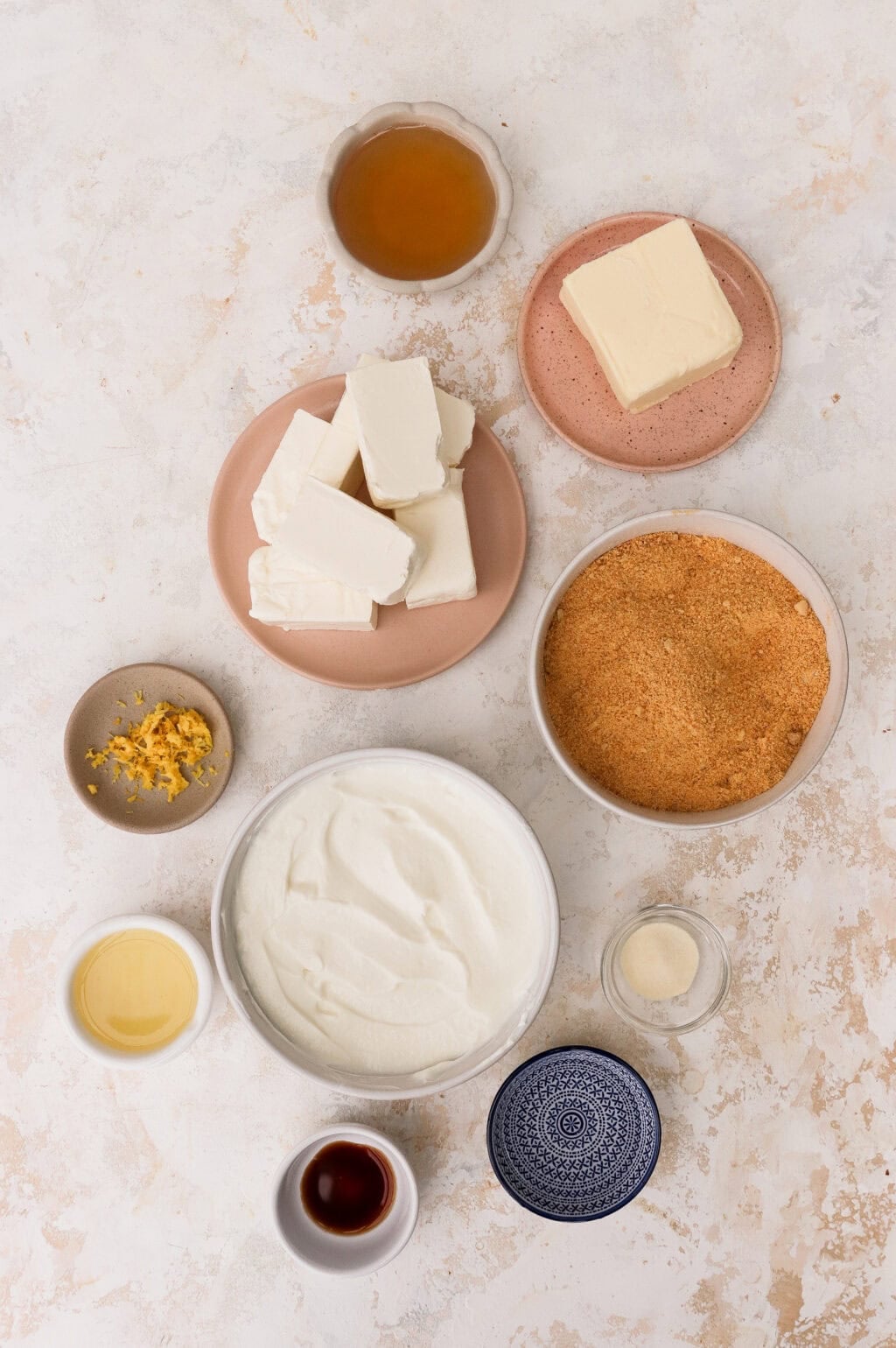 Overhead ingredients flat lay for no bake cheesecake — cream cheese blocks, bowl of whipped cream, graham cracker crumbs, butter, vanilla extract, and lemon on marble surface