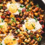 One-pan camping breakfast skillet with eggs, golden potatoes, red and green peppers, and sausage in a rustic cast iron pan over a campfire, steam rising, outdoor morning light