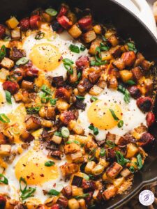 One-pan camping breakfast skillet with eggs, golden potatoes, red and green peppers, and sausage in a rustic cast iron pan over a campfire, steam rising, outdoor morning light