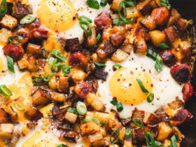 One-pan camping breakfast skillet with eggs, golden potatoes, red and green peppers, and sausage in a rustic cast iron pan over a campfire, steam rising, outdoor morning light