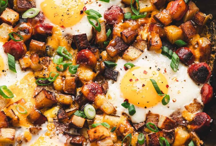 One-pan camping breakfast skillet with eggs, golden potatoes, red and green peppers, and sausage in a rustic cast iron pan over a campfire, steam rising, outdoor morning light