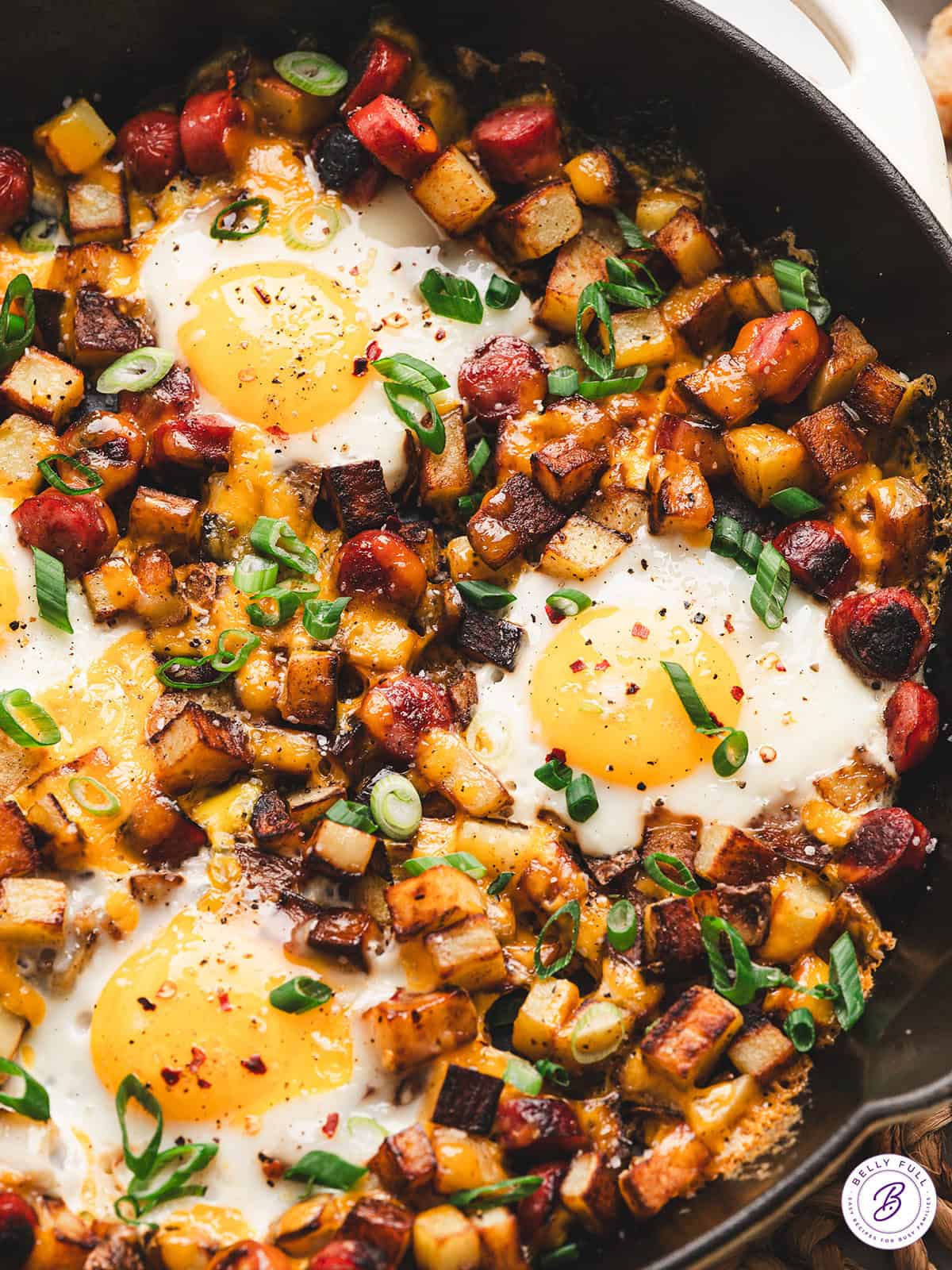 One-pan camping breakfast skillet with eggs, golden potatoes, red and green peppers, and sausage in a rustic cast iron pan over a campfire, steam rising, outdoor morning light