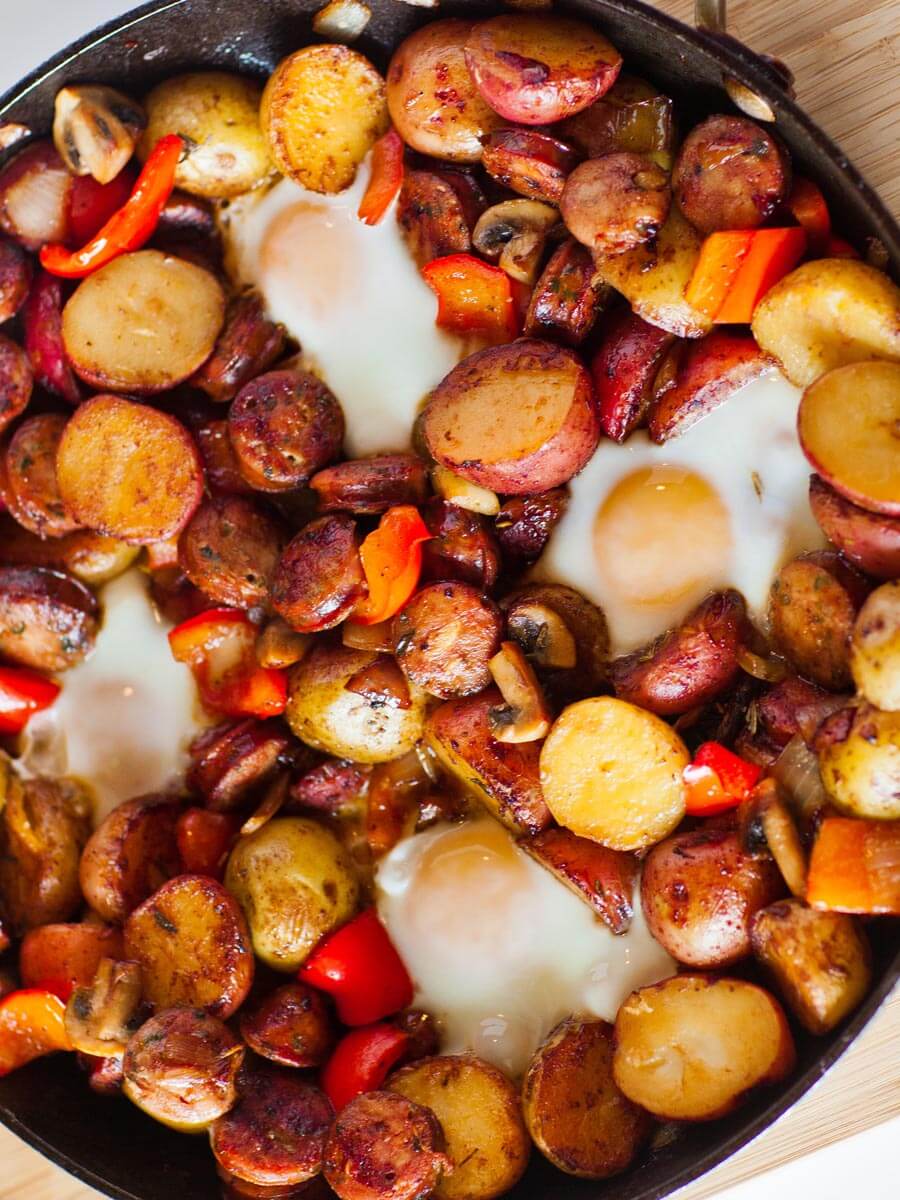 Overhead flat-lay of camping breakfast skillet ingredients — diced potatoes, sliced colorful peppers, raw sausage links, eggs, garlic, smoked paprika, and olive oil arranged on a rustic wooden camp table