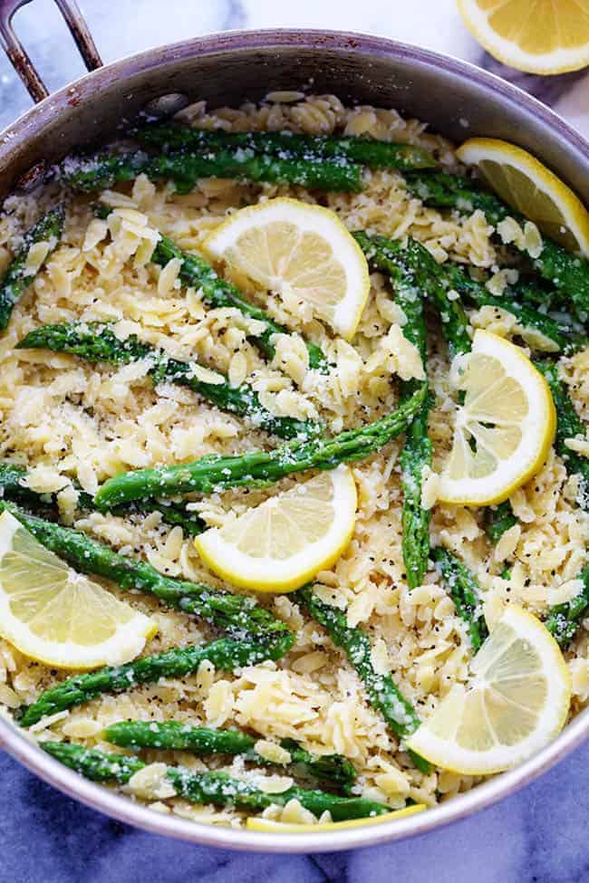 Overhead flat-lay of orzo, asparagus spears, parmesan block, garlic cloves, shallot, broth carton, and lemon arranged on a white marble surface