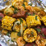 Shrimp boil foil packet opened on a plate showing plump shrimp, smoked sausage slices, golden corn, and baby potatoes garnished with parsley and lemon wedges