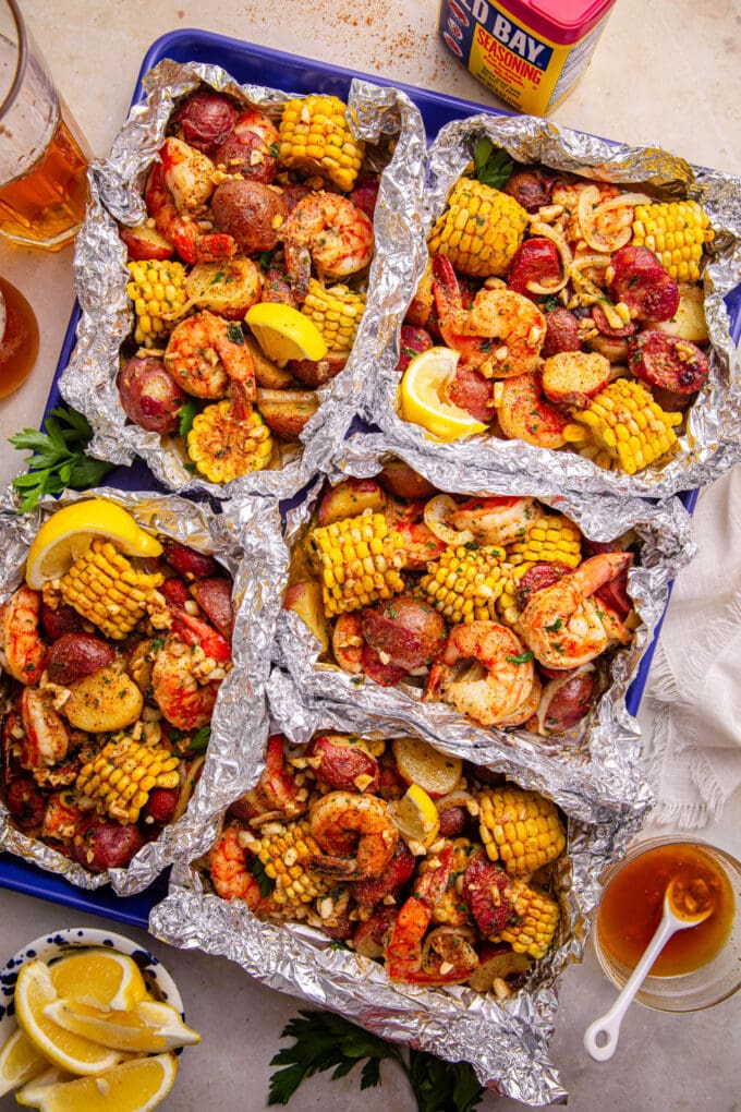 Flat lay of shrimp boil foil packet ingredients including raw shrimp, sliced andouille sausage, baby potatoes, corn rounds, butter pats, lemon slices, and Old Bay seasoning on a wooden surface