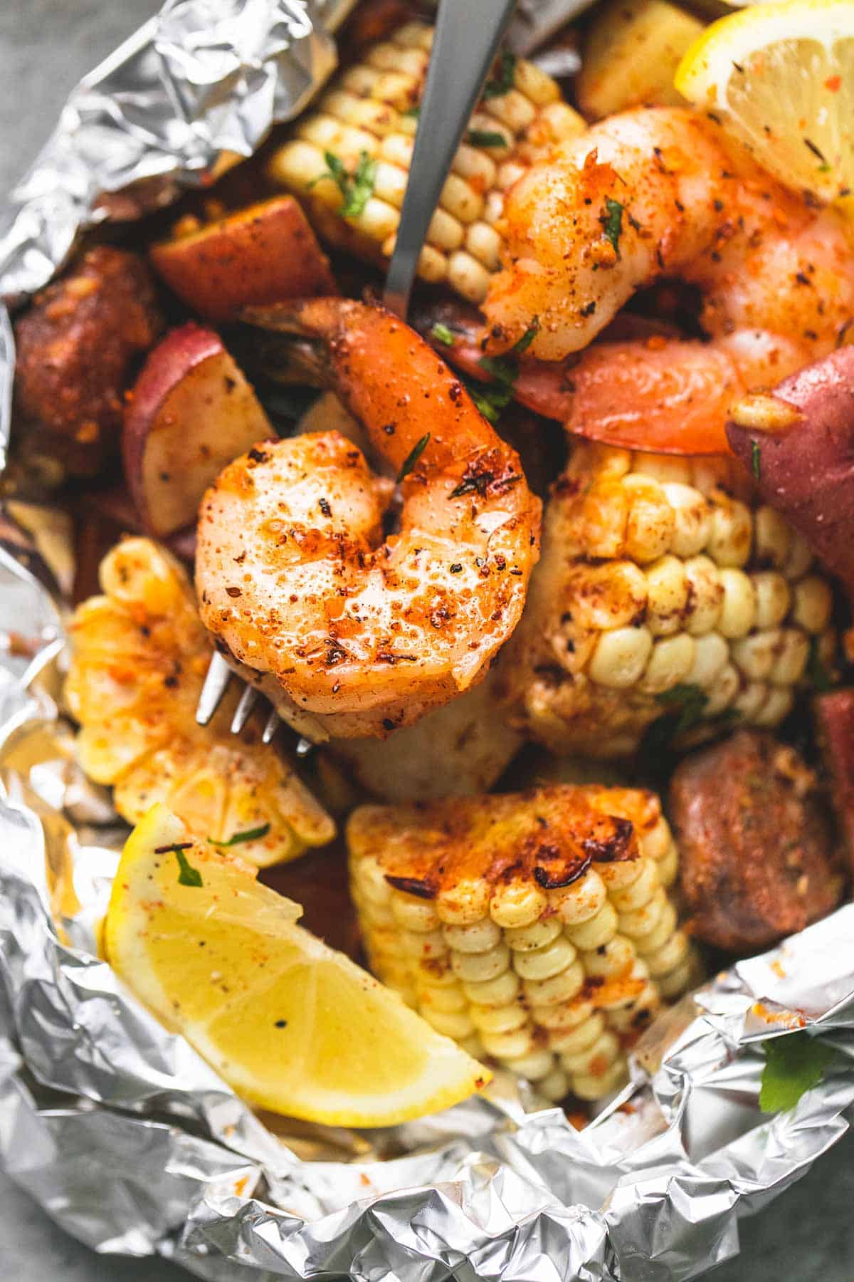 Close-up of an opened shrimp boil foil packet with juicy shrimp, charred corn, and smoked sausage served with crusty bread and lemon wedges on a rustic outdoor table