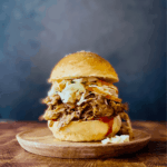 Slow cooker pulled pork served on a toasted bun with coleslaw and pickles, beautifully plated on a rustic wooden board