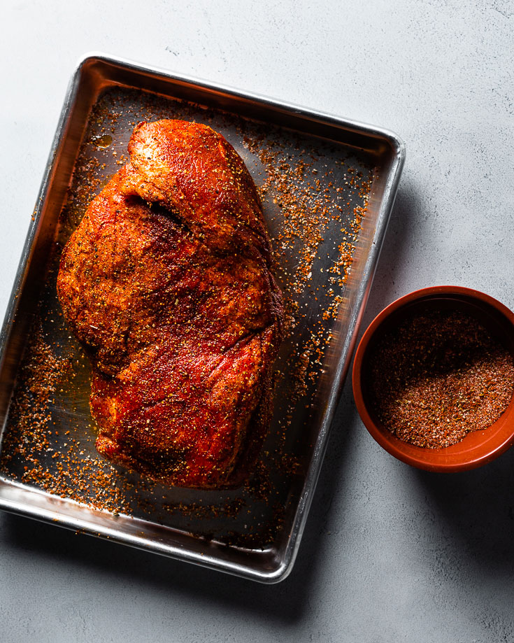 Pork shoulder coated in dry spice rub next to small bowls of individual spices and apple cider vinegar on a clean kitchen counter