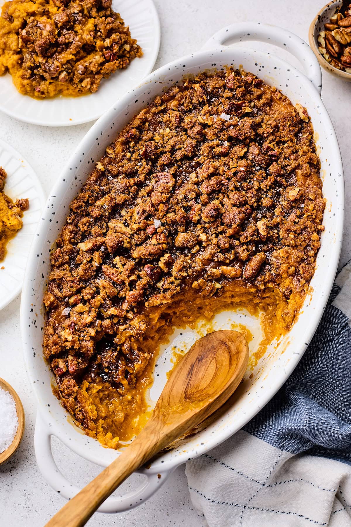 A flat lay ingredients shot showing peeled sweet potatoes, pecans, maple syrup, butter, cinnamon sticks, and vanilla extract arranged on a marble surface