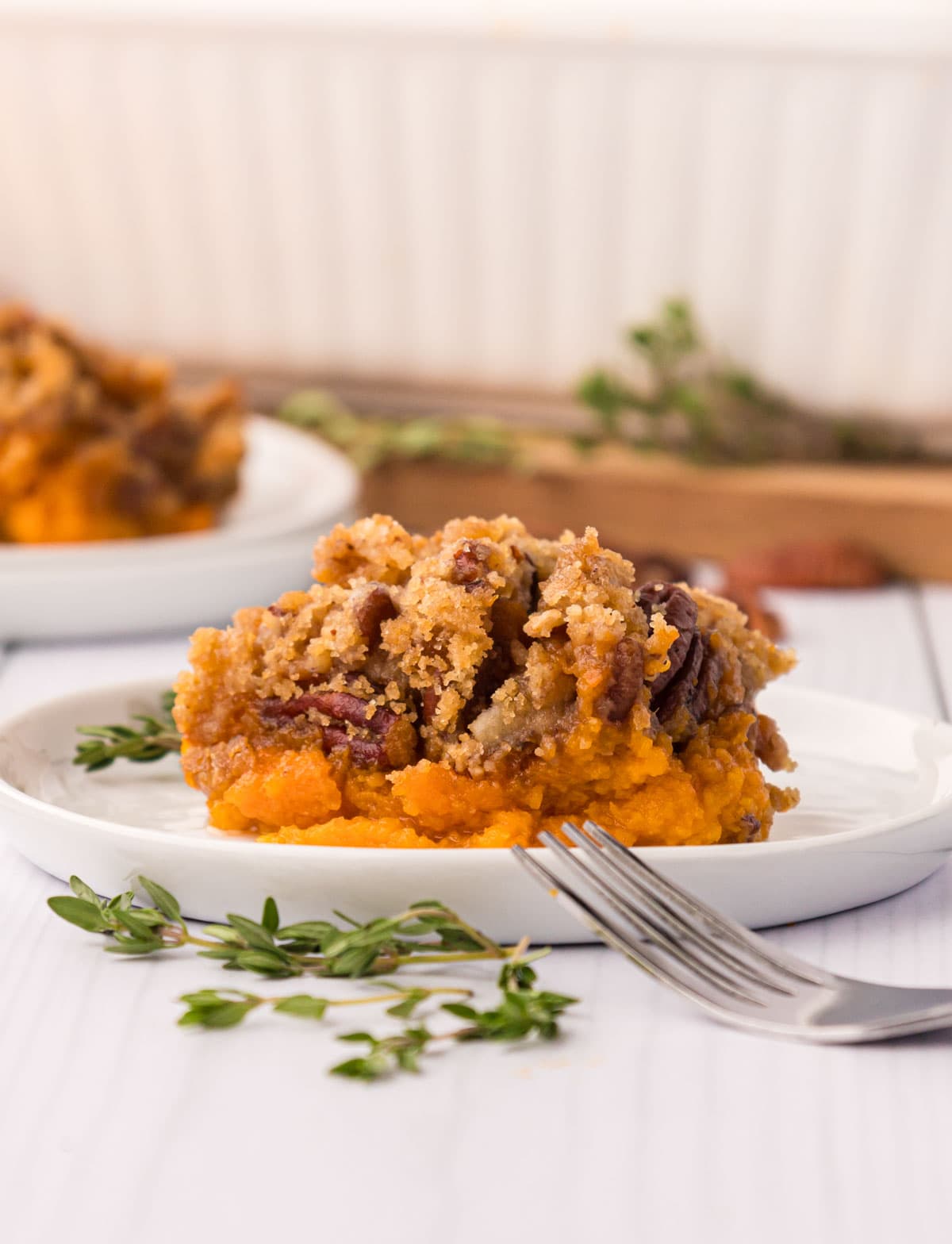 A close-up serving of sweet potato casserole on a white plate showing the creamy orange filling and crunchy golden pecan topping, with a fork resting on the plate