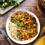 A deep bowl of Sweet Potato Peanut Stew garnished with fresh cilantro, a swirl of peanut butter, sliced chili, and served over fluffy white rice on a rustic wooden table