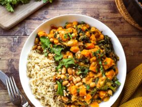 A deep bowl of Sweet Potato Peanut Stew garnished with fresh cilantro, a swirl of peanut butter, sliced chili, and served over fluffy white rice on a rustic wooden table