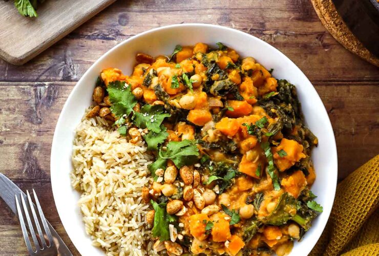 A deep bowl of Sweet Potato Peanut Stew garnished with fresh cilantro, a swirl of peanut butter, sliced chili, and served over fluffy white rice on a rustic wooden table