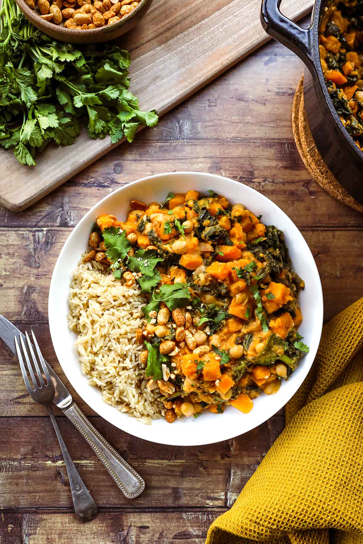 A deep bowl of Sweet Potato Peanut Stew garnished with fresh cilantro, a swirl of peanut butter, sliced chili, and served over fluffy white rice on a rustic wooden table