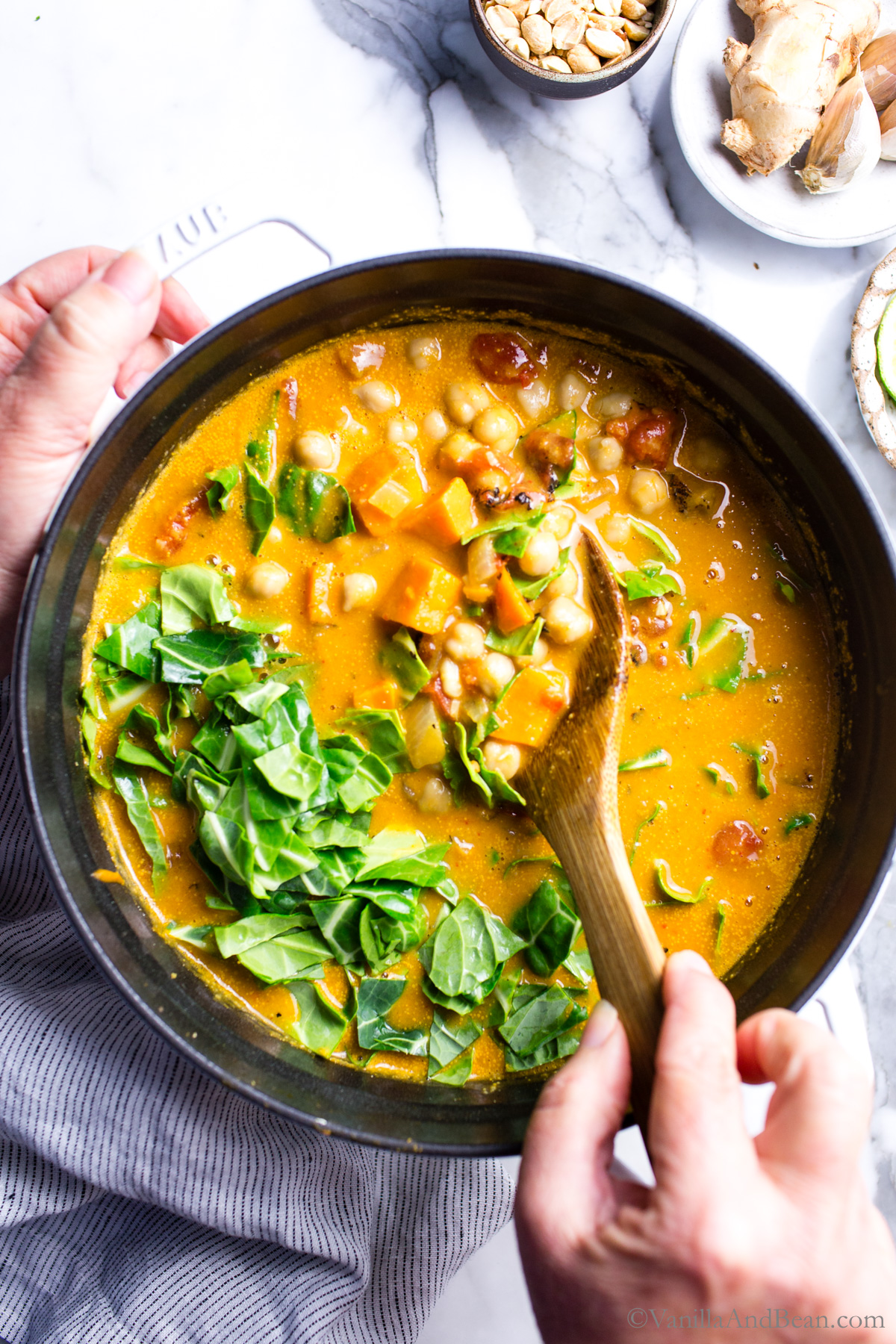 A close-up of a spoonful of Sweet Potato Peanut Stew being lifted from a bowl, showing chunks of orange sweet potato, chickpeas in a creamy peanut broth, with wilted greens and garnished with crushed peanuts and cilantro