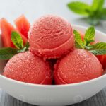 Watermelon sorbet scooped into a white ceramic bowl garnished with fresh mint and a thin watermelon slice, shot overhead on a bright white marble surface with a linen napkin and silver spoon
