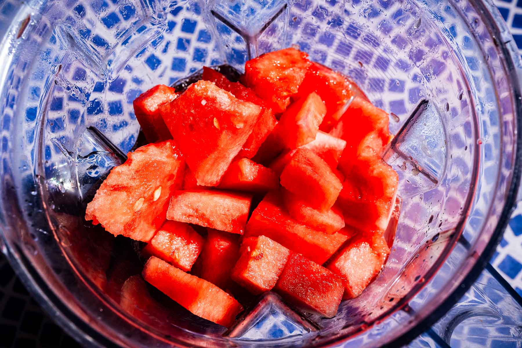Cubed fresh watermelon pieces spread on a parchment-lined baking sheet ready to go into the freezer, with a lime, honey jar, and sea salt arranged alongside on a wooden surface