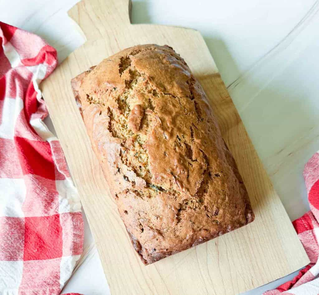 Zucchini Bread — Moist Cinnamon Spiced Quick Bread You'll Make All Summer 26 Sliced zucchini bread loaf on a wooden cutting board with a pat of butter melting on the top slice, warm natural light, fresh zucchini in the background