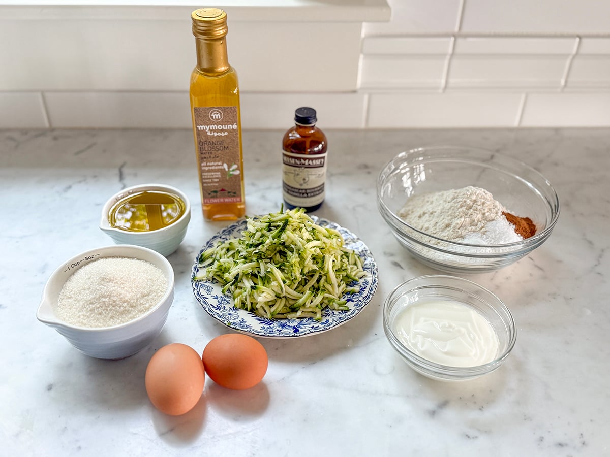 Zucchini Bread — Moist Cinnamon Spiced Quick Bread You'll Make All Summer 27 Overhead flat lay of zucchini bread ingredients — grated zucchini in a bowl, cinnamon sticks, eggs, flour, oil, and brown sugar on a white marble surface
